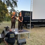Zwei Gebärdensprachdolmetscherinnen stehen neben der Leinwand und ein Eventtechniker prüft die Technik.