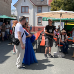Menschen stehen und sitzen bei der Veranstaltung "Bandmeeting" mit Tischen, Bänken und Sonnenschirmen vor der Bühne.